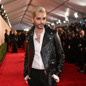 Bill Kaulitz at a glitzy awards show. Black leather jacket and white shirt. Poses on red carpet. Crowded and glamorous atmosphere.