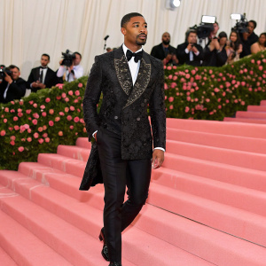 Michael B. Jordan walking the red carpet at the Met Gala. Wearing a stunning black tuxedo with intricate detailing. Pink steps and white curtains in the background.