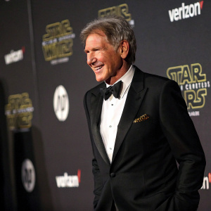 Harrison Ford in sharp black suit, bow-tie and slicked back hair, smiles warmly at event. Dark, dramatic backdrop with company logos.