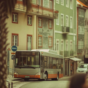 A Van Hool A330 city bus, gray and orange, parked on a bustling street with unique architecture. Buildings with diverse facades and balconies create a charming European scene. Ads and a bus stop sign are visible in the background.