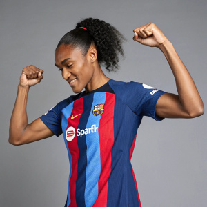 Salma Paralluelo, an athlete, posing confidently in a Nike soccer jersey. The jersey has a mix of blue and red with white accents. Flexing muscles in a grey studio setting. High resolution photo.