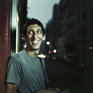 Angel Di Maria, in a casual gray t-shirt, smiling broadly. Shot from a distance, blurred cityscape in the background.