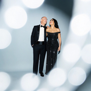 Lauren Sanchez and Jeff Bezos attending the Met Gala. Sanchez, of Hispanic descent, wearing a black sequined gown. Bezos in a black tuxedo. Blurred background of colorful lights.