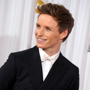 Eddie Redmayne, in a black suit and white shirt, at the Golden Globe Awards. He looks charming with his neat hair and warm smile. The background features a plain white curtain.