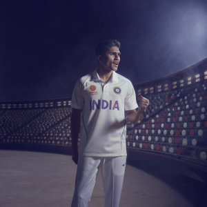 Shubman Gill, young Indian cricketer, standing against a vibrant stadium backdrop. He is wearing an athletic white jersey with 'India' in bold letters and an embedded sponsor logo. His short hair and youthful look are prominent.