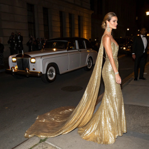 Rosie Huntington-Whiteley at the Met Gala. Wearing a shimmering gold gown with dramatic train, standing on a dimly lit street with vintage Rolls-Royce in background.
