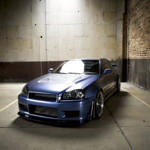A high-performance Nissan GTR parked in an urban alleyway. The car has a blue metallic paint job with a sleek, low-slung design. Distinctive front grille and hood ornament visible. Dramatic lighting creates a flat, textured surface effect.