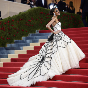 Winnie Harlow, posing on the stairs of the Met Gala. Wearing a white and black gown with intricate layers. Formal attire, red carpet backdrop.