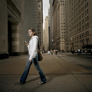 Emma Watson in a casual outfit, blue jeans, and a white hoodie. She is walking through a bustling city street with tall skyscrapers in the background.