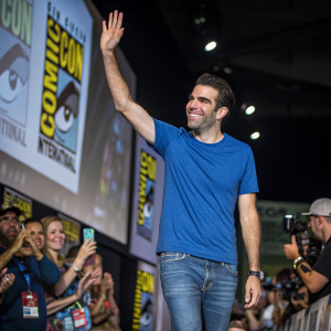 Zachary Quinto at Comic-Con, wearing a blue t-shirt and jeans. Surrounded by fans, smiling and waving