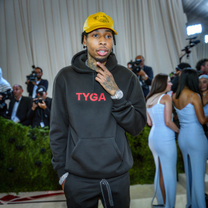 Tyga, sporting a yellow cap and face tattoos, at the Met Gala. Wearing a black hoodie with red text. Amidst a sea of celebrities and paparazzi.