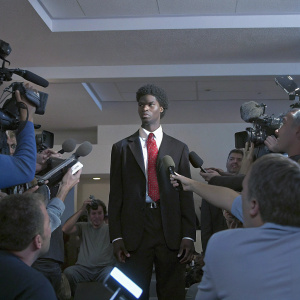 Shai Gilgeous-Alexander at a press conference. Wearing a black blazer and red tie. Crowded room with flashing cameras
