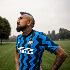 Arturo Vidal, the lion-hearted player for Inter Milan, posing confidently. His mane of hair and rugged beard evoke strength. The grassy expanse of a quiet park stretches behind him, a stark contrast to the vibrant blue and black jersey with the sponsor logo.