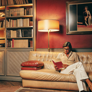 Helmut Newton, sitting on a vintage leather sofa in a cozy, dimly lit study. The room filled with books and photographs.