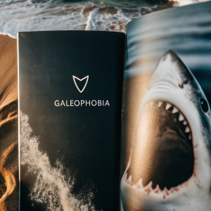 Page dedicated to galeophobia featuring a dramatic photo of a shark. Right side has an artistic logo of a shark tooth. Left side includes sleek text detailing the phobia. Modern, ocean-themed design.