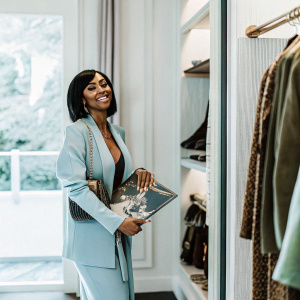 Keri Hilson, an African American woman with a trendy haircut, smiling while holding her album cover. She's in a luxurious boutique filled with designer clothing racks.