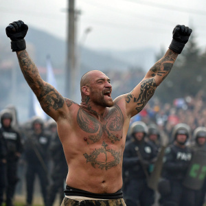 Burly, bald warrior with tattoos, raising his arms in triumph. Crowded background, blurred guards and onlookers.