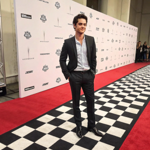 Dylan O'Brien at award show, standing confidently on red carpet. Black and white checkered floor. Minimalist backdrop with sponsor logos