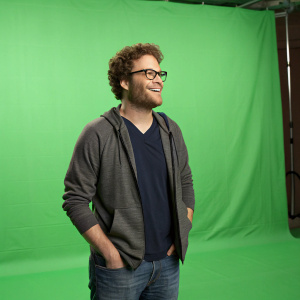 Seth Rogen, curly hair, glasses, in casual attire. Green screen background, standing in studio.