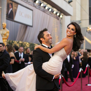Daniel Sadek lifts Nadia Bjorlin into his arms with a smile, standing before a bustling crowd at a glamorous awards ceremony.