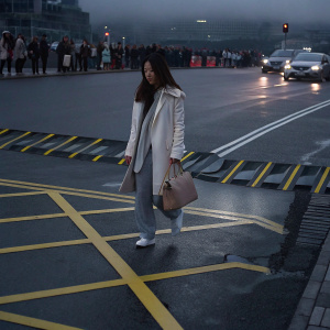 Myleene Klass outside a bustling cityscape. Wearing a white coat and gray sweatpants. Tan purse in hand, walking past yellow caution lines on a paved street.