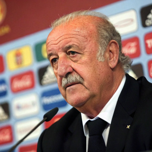 Vicente del Bosque, former football manager, wearing a black suit and tie, addressing the press. Vibrant background with multiple logos. Close-up shot focusing on his mustachioed face and upper body. Even lighting throughout the scene.