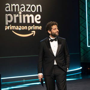 Joe Lewis, Hispanic male with beard and curly hair, standing on a dimly lit stage at the Amazon Prime Awards. Wearing a classic tuxedo, white shirt, and black bow tie. The backdrop is sleek and modern with repeated Amazon Prime branding and hashtag #AmazonPrime.
