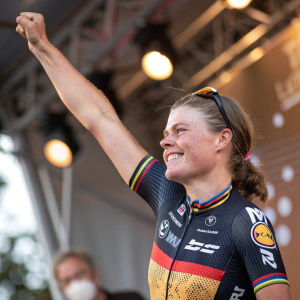 Lotte Kopecky in a victorious pose. Celebrating with one arm raised. Wearing a sleeveless jersey with various sponsors' logos. Joyful expression with a mask on.