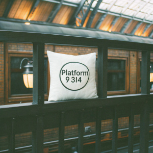 Photograph of a white pillow featuring the Platform 9 3/4 logo. Set against a backdrop resembling the interior of a cozy, wooden train station. Part of the Harry Potter series theme.