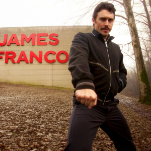 James Franco, a white male with mustache, striking a confident pose. Dressed in black zip-up jacket with zipper and stripe visible, paired with dark pants. Set against a vivid yellow backdrop adorned with bold red text. Close-up, high-resolution shot highlighting Franco.