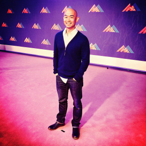 Tai Trang, Asian male, bald head, smiling on the award show red carpet. Wears a navy sweater over a crisp white shirt, paired with black boots and dark jeans. Vibrant backdrop featuring a mix of colorful logos and graphics.