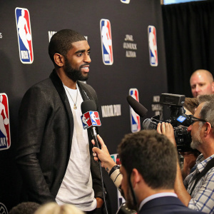 Kyrie Irving in a press conference. Surrounded by journalists. Background is black, with NBA logos visible