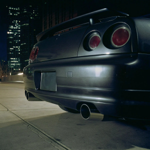 Photograph of a Nissan Skyline GTR R34 in matte black. Detailed shot of the rear bumper and exhaust. Cityscape background