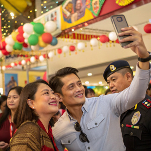 Robin Padilla taking a selfie with a woman and a security officer. Festive event with decorations and a lively crowd.