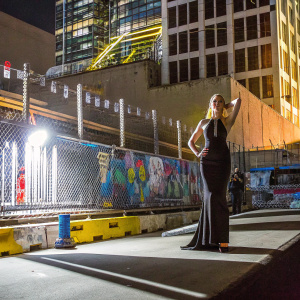 Sophie Turner, in a sleek black dress, posing in front of an urban graffiti wall. Dramatic evening light, casting sharp shadows