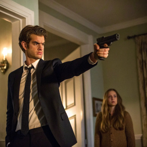 Andrew Garfield, in dark suit and formal tie, pointing a gun in a dimly lit room. Long-haired woman in background. Both characters are in a tense standoff.