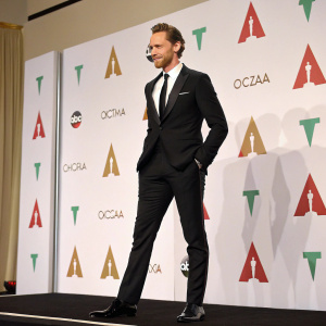 Tom Hiddleston, standing confidently on the stage at the Oscar Awards. Wearing black suit, white shirt, and black tie. White backdrop with logos and red and green color scheme.