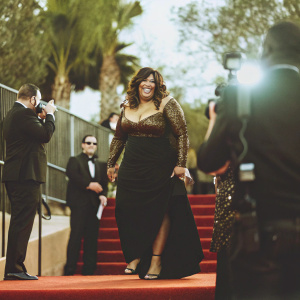 Kym Whitley, African-American actress, with radiant smile and elegant demeanor. Black gown with gold accents, stilettos. Event: Grammy Awards. Red carpet, flashing cameras.