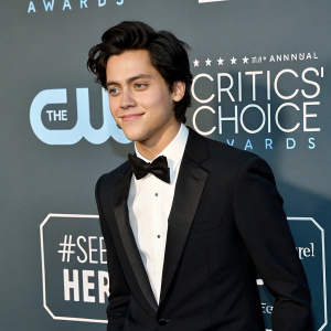 Cole Sprouse, dressed in a classic black tuxedo with a crisp white shirt and bow tie, at the annual Critics' Choice Awards. A backdrop with event branding