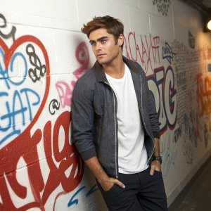 Zac Efron, casually dressed, standing in front of a bold graffiti wall covered in text and graphics. Indoor studio setting, close-up shot