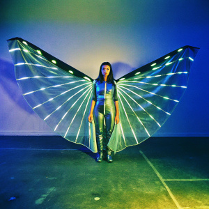 Larissa Andrade, wearing a futuristic winged outfit, standing alone in a dimly lit studio. Serious expression
