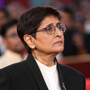 Close-up of Kiran Bedi, former IPS officer, wearing a black suit and white blouse. Serious demeanor. Background: blurred audience at an awards ceremony.