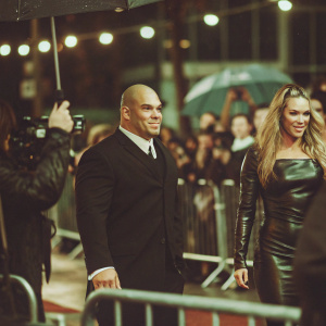 Tito Ortiz and Amber Nichole Miller at a bustling New York City premiere event. Tito, bald and in a black suit, stands beside Amber in a black leather dress, both basking in the spotlight of flashing cameras