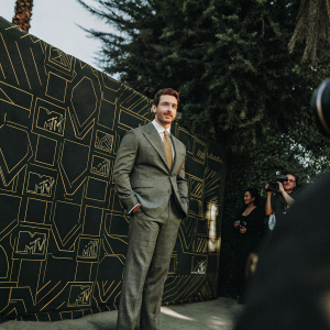 Miles Teller at the MTV Movie Awards. Grey suit with gold tie. Vibrant backdrop with neon MTV logos