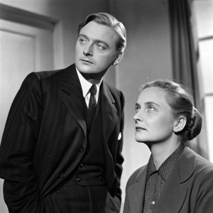 Oskar Werner and Gertrud Kueckelmann captured in a vintage studio shot. Oskar, in a sleek suit, stands beside Gertrud, who sits with a serious expression and hair in a neat bun.