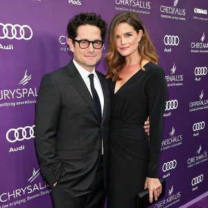 J.J. Abrams, in a dark suit with glasses, and Katie McGrath, in a sleek black dress with wavy hair, pose together at the 12th Annual Chrysalis Butterfly Ball. Purple backdrop with logos and sponsor names behind them.