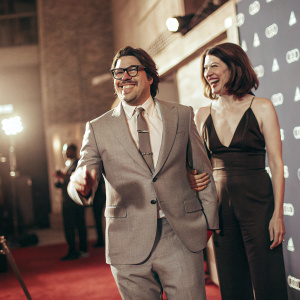 Rich Sommer and Virginia Donohoe at a Hollywood party. Rich, with Hispanic heritage, in a classic gray suit, tie, and glasses. Virginia, in a sleek black jumpsuit. Behind them, an eye-catching blue backdrop adorned with Audi logos. Both beaming with joy.