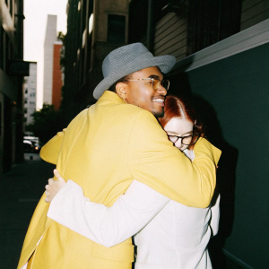 Ne-Yo, a man with warm smile and gray fedora hat, hugged by a woman with red hair. Another woman in glasses stands nearby. Scene takes place on a sunny street with urban buildings.