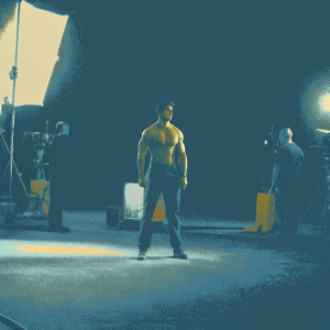 Robbie Amell, shirtless, on set of action movie. Bright lights and crew visible in background.