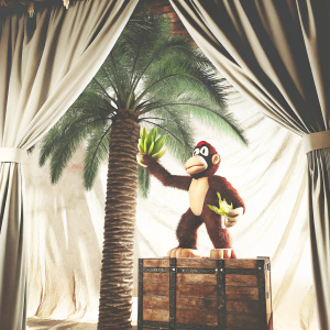 A digital 3D rendering of Diddy Kong from the Donkey Kong series. Diddy Kong stands dynamically on a wooden crate with a palm tree in the background. The scene is set against a soft, flowing fabric backdrop. The overall color palette is warm and varied with rich textures.